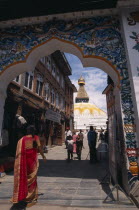 A woman wearing a red sari walking through ornate Gateway towards Bodhnath Buddhist Stupa.