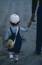 Young child seen from behind  wearing white cap and short trousers holding on to finger of adult  partly seen  at side.
