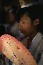 Portrait of young shrine maiden employed to welcome visitors  supervise offerings and perform sacred dances for the Shinto gods or kami.  Head and shoulders  three-quarter portrait to left  carrying painted fan.