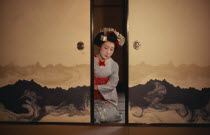Portrait of Someiyu  a maiko or trainee geisha kneeling in tightly bound kimono between partly open screens with traditional make-up and hair piece in readiness for her evening appointments.