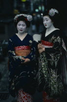 Two maiko or apprentice geisha with white powdered faces and red painted lips wearing richly embroidered kimonos and decorative hair pieces.
