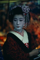 Three-quarter portrait of sixteen year old Someiyu  a maiko or apprentice geisha with white powdered face and red painted lips wearing richly patterned kimono and hair decoration.
