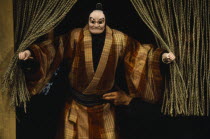 Bunraku puppet male character appearing from behind curtains with puppeteer partly seen at side.