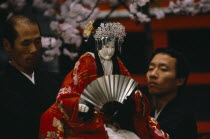 Bunraku puppet female character with puppeteers.   Puppet is half life size  dressed in red  embroidered kimono and holding fan.