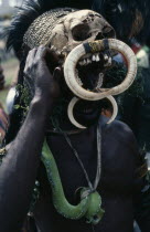 Head and shoulders portrait of man wearing traditional head-dress of human skull adorned with pig tusks and a live snake as a necklace.  Masks and face painting are used to proclaim personal prestige...