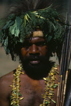 Head and shoulders portrait of man wearing head-dress made from leaves and vegetation with fur band around forehead and double strand necklace of leaves.
