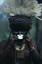 Portrait of man wearing traditional dress and jewellery  and elaborate white feather and fur trimmed head-dress  with white disc in nose and black and white face paint.