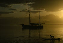 Sunset reflected in still expanse of water with silhouetted sail boat at anchor and children playing in the water in foreground.