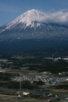 Snow capped peak of Mount Fuji rising up behind houses and agricultural land.