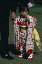 Little girl holding the hand of a part seen guest at Shinto wedding ceremony wearing traditional dress printed with red and white squares and flowers and pale yellow sash.