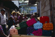 Market scene with wool stall piled with brightly coloured bundles of wool and crowd of customers wearing traditional dress.