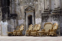 Street scene with line of wicker chairs and tables outside building with crumbling  discoloured plaster wall.