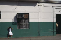 Street scene with little girl in white outside building painted dark green and white.  Metal window grill casting shadow above and man standing in doorway on right.