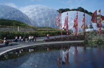 Eden Project.  General view over the Humid tropics Biome exterior with line of coloured flags and visitors walking beside lake.