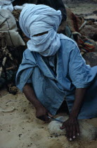 Taureg man sharpening tool on rock  wearing blue taguelmoust covering face and head except for eyes. In Tuareg societies men are veiled rather than women.