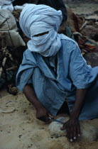 Taureg man sharpening tool on rock  wearing blue taguelmoust covering face and head except for eyes. In Tuareg societies men are veiled rather than women.