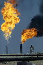 Flaming gas on oil field with man standing behind pipe in foreground.