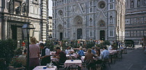 Cafe in front of the Neo-Gothic marble facade of the Duomo with people sitting at outside tables having coffee and reading guide books.