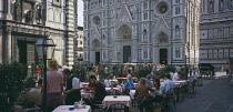 Cafe in front of the Neo-Gothic marble facade of the Duomo with people sitting at outside tables having coffee and reading guide books.