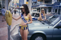Meter Maids in bikinis feeding parking meters with coins.