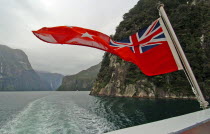 SOUTHLAND  A NEW ZEALND ENSIGN FLAG WAVES ON THE REAR OF REAL JOURNEYS CRUISE BOAT THE MILFORD MARINER AS IT TRAVELS ALONG MILFORD SOUND IN NEW ZEALANDS FJORDLAND AREAAntipodean Oceania