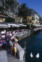 Limone Sur Garda.  Busy lakeside cafe with people sitting at tables under awning.  Woman and child feeding swans in the foreground.