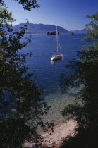 Yacht anchored on lake framed by trees in foreground.  Stretch of pebble shore and clear  shallow water below.
