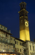 Torre dei Lamberti clock tower in the Piazza Erbe illuminated at night part seen behind faded building facades  statue and lights and umbrellas of cafe bar.