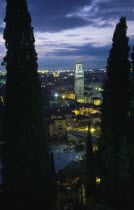 Cityscape illuminated at night showing tiled rooftops  Duomo bell tower and bridge over the Adige River part framed by tall coniferous trees.  Windswept evening sky.