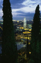 Cityscape illuminated at night showing tiled rooftops  Duomo bell tower and bridge over the Adige River part framed by tall coniferous trees.  Windswept evening sky.