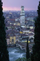 Cityscape at sunset showing tiled rooftops  Duomo bell tower and bridge over the Adige River part framed by tall coniferous trees.  Pale pink/purple sky and city lights.