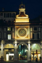 Astronomical clock in Piazza della Loggia illuminated at night with brightly lit shop windows framed by archways of colonnade below.  People  street lamps and light trail.