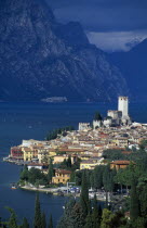 Malcesine.  Tiled rooftops and pastel coloured buildings of town and castle on shore of lake with mountains beyond.