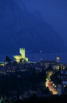 Malcesine.  Tiled rooftops of town and castle illuminated at night with lake beyond.  Coloured light trails from cars on road in foreground.