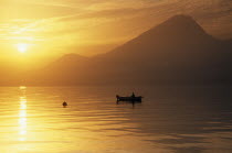 Fishing boat on lake at sunset in golden light with mountain peak in haze of sunshine behind.
