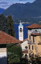 Stresa.  Overhanging red tiled rooftops of town buildings with church bell and clock tower in centre.  View across lake towards mountain behind.