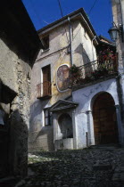 Narrow cobbled street with fresco decoration on exterior wall of building and drinking fountain below beside arched doorway with balcony and flowers above.