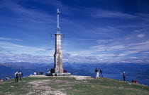 Tourists at summit of Monte Mottarone marked by tower and cross overlooking Lake Maggiore and the surrounding area.