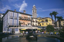 Pallanza.  Bar and cafe with people sitting at outside tables in  square with central circular fountain.  Pastel coloured building facades and bell tower part seen above