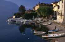 Sala Comacina.  Fishing village with motor boats moored against wooden jettys extending from pebble beach overlooked by pastel coloured facades of houses and cafe
