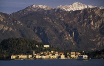 Ballagio. View across Lake Como towards distant town of Ballagio situated at foot of tree covered hillside with mountain backdrop. Car ferry crossing lake in foreground.