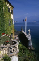 Villa Balbianello.  Part view of ivy and virginia creeper covered facade  stone balcony with pots of red geraniums and wall with statues and flags. View over lake beyond.