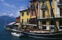 Yellow  white and terracotta painted facades of waterside buildings with moored yacht and motor boats in foreground.  People at cafe at end looking out across lake