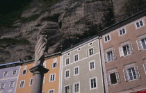 Pastel coloured houses on Gstatiengasse built into the Monchsberg beneath the fortress with statue on plinth in foreground.