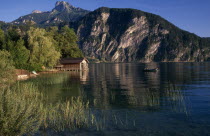 Small motor boat approaching boat house on shore of Monsee Lake with mountain backdrop reflected in rippled surface  reeds and willow trees.