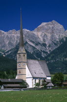 Austria, Salzburg, Maria Alm am Steinernen Meer Parish church exterior with long  narrow spire with Hochkonig mountain range behind.  Near Saalfelden.