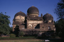 Unidentified old building in Islamic architectural style with three domes set in gardens or green space with person asleep beside bicycle in foreground.