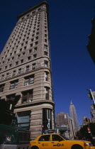 Angled view of the Flatiron Building from 23rd Street.  Traffic including yellow taxi cab below and Empire State Building beyond.Steel framed  Beaux-Arts skyscraper designed by architect Daniel Burn...