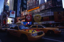 Times Square.  Yellow taxi cabs on street lined with skyscraper buildings and illuminated advertising in low light.