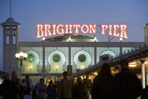 Entrance to the Brighton Pier at dusk showing illuminated sign and people on the walkway.
